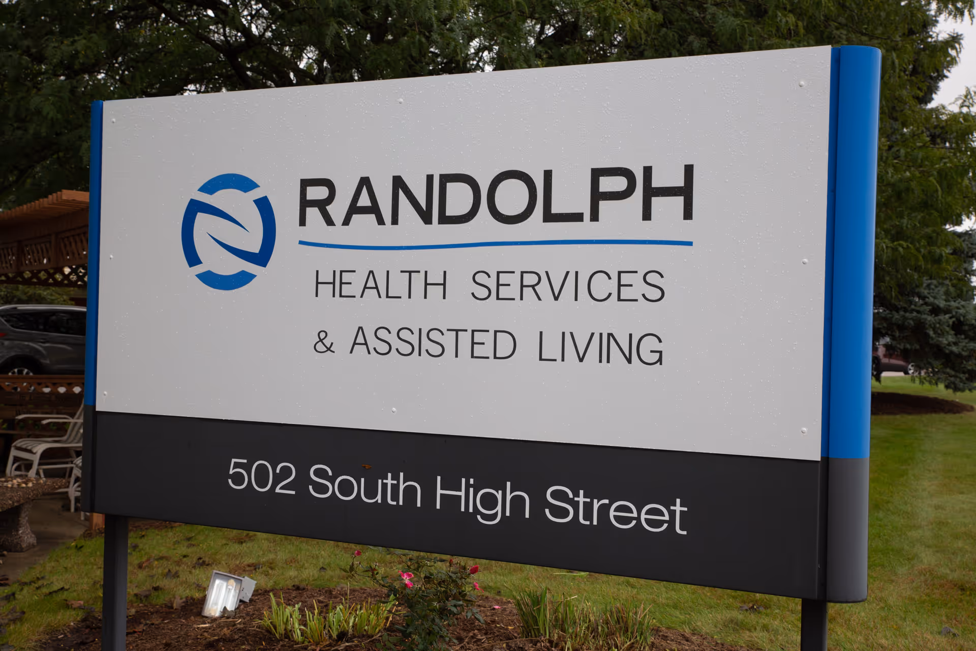 Outdoor sign for Randolph Health Services & Assisted Living located at 502 South High Street, surrounded by grass, plants, and trees.