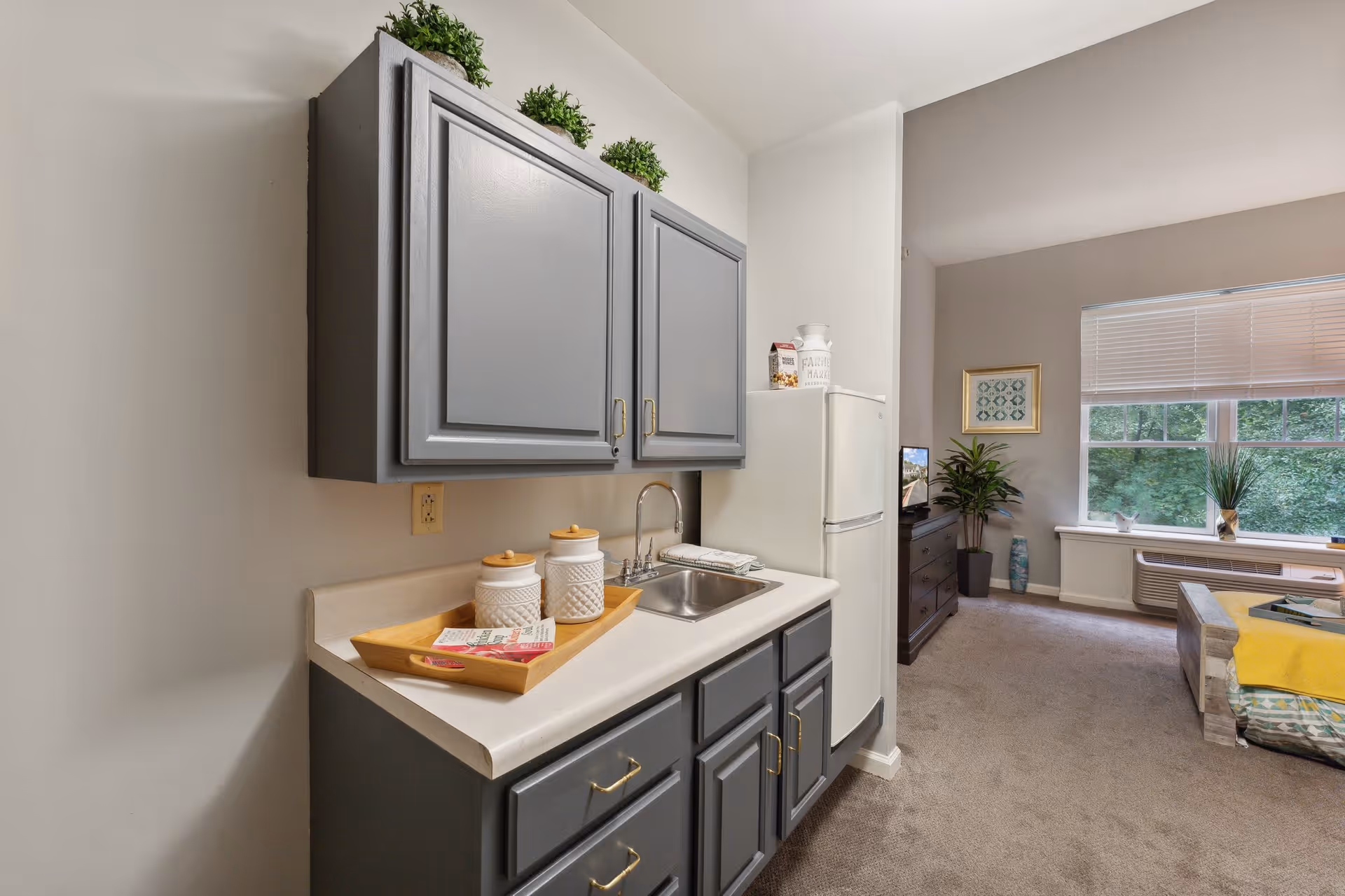 A small kitchenette area with gray cabinets, a white countertop, a stainless steel sink, and a white refrigerator. On the countertop, there are three white canisters on a wooden tray and a folded towel next to the sink. The kitchenette opens into a living area with a window, a potted plant, a dresser with a TV, and a couch with a yellow blanket.