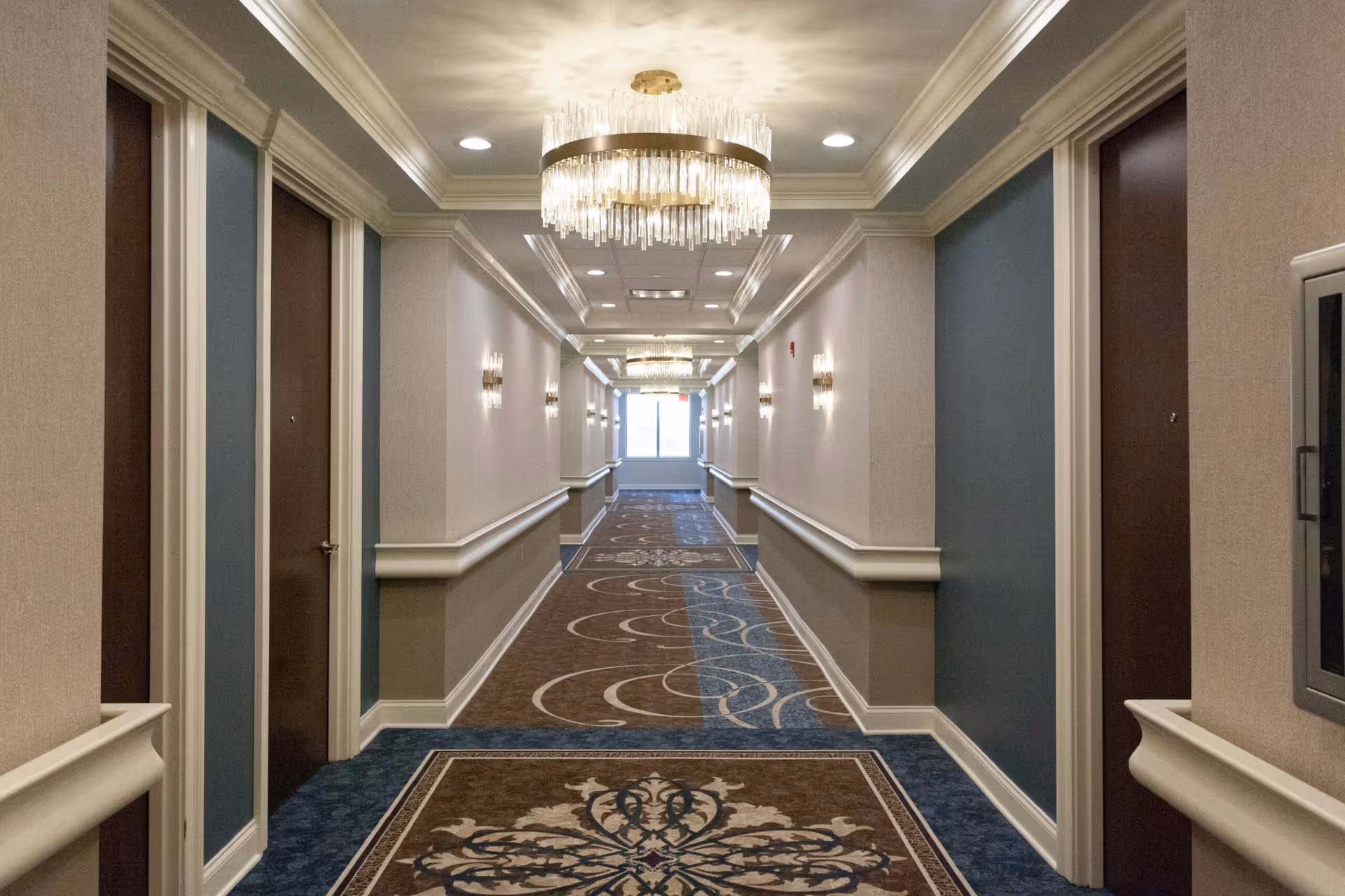 A long, well-lit hallway in a senior living facility with patterned carpet, beige and blue walls, multiple closed brown doors on both sides, decorative wall sconces, and two large crystal chandeliers hanging from the ceiling.