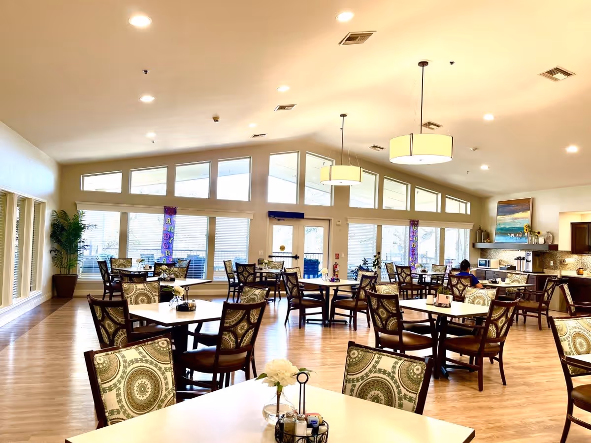 A spacious dining room with multiple tables and chairs arranged neatly. The room has large windows allowing natural light to fill the space. There are decorative plants and banners near the windows. The ceiling has modern hanging light fixtures, and there is a kitchenette area with cabinets, a microwave, and a coffee machine in the background. One person is seated at a table near the kitchenette.
