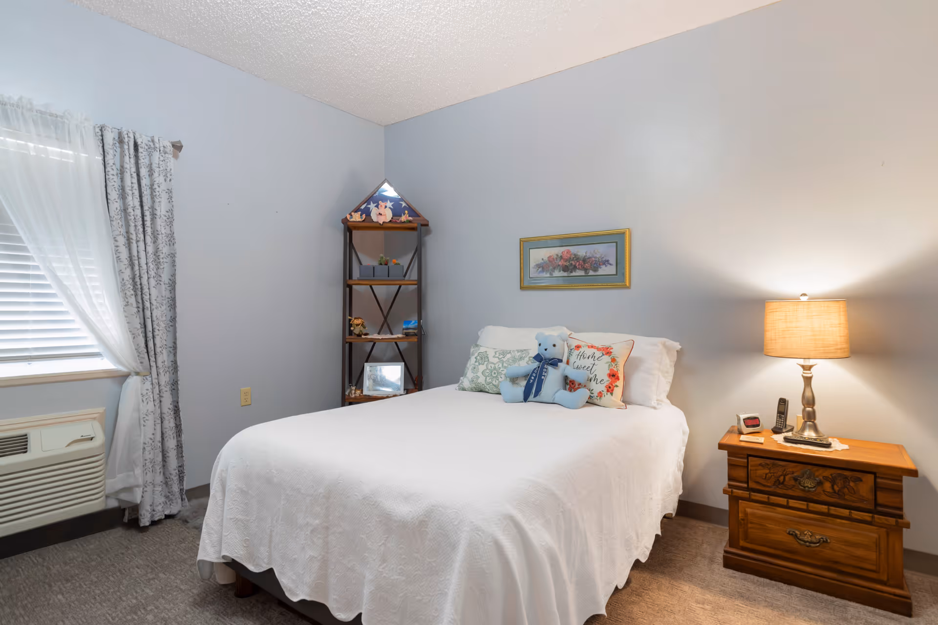 A cozy bedroom with a neatly made bed covered in a white quilt, adorned with decorative pillows including a blue teddy bear. To the right of the bed is a wooden nightstand with a lamp, a phone, and a clock. A framed floral picture hangs on the light blue wall above the bed. To the left, there is a window with white blinds and sheer curtains, and a small air conditioning unit below it. A corner shelf with decorative items is placed against the wall.