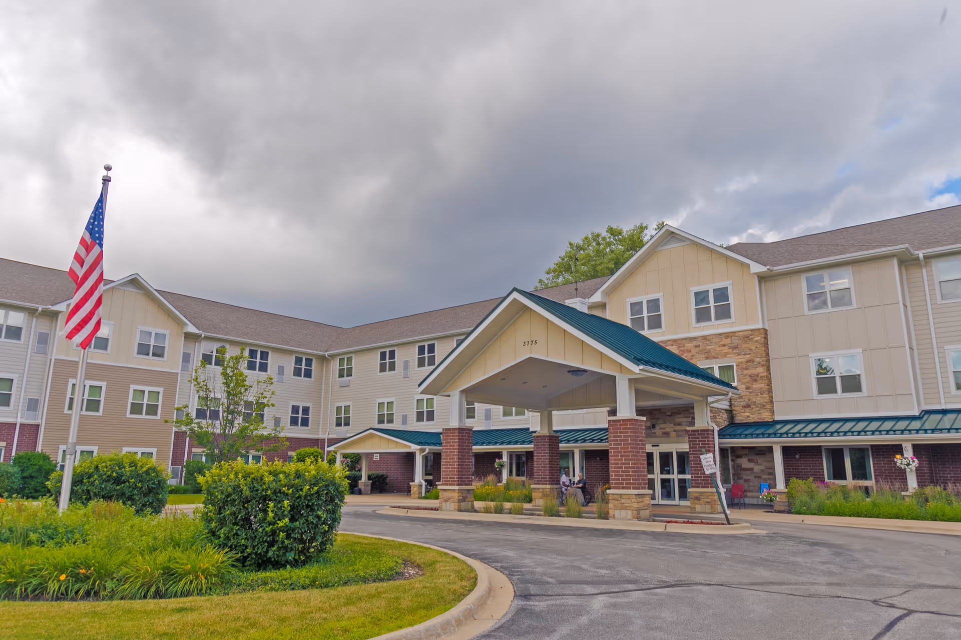Exterior view of Heritage Woods of Gurnee, a multi-story senior living facility with beige and brick facade, a covered entrance supported by brick pillars, an American flag on a flagpole, and landscaped greenery in front under a cloudy sky.