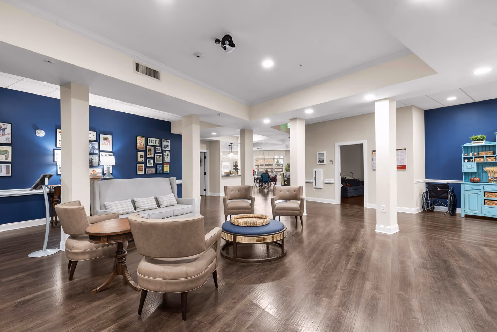 A spacious and well-lit common area in a senior living facility with wooden flooring, beige and blue walls, and comfortable seating including a gray sofa, beige armchairs, and a round coffee table. The walls are decorated with framed pictures and a blue cabinet with baskets and plants is visible. A wheelchair is parked near the cabinet, and a dining area can be seen in the background.