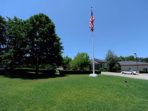 A grassy lawn with a tall flagpole displaying the American flag in the center. There are several trees on the left side and a building with a driveway and parked car on the right under a clear blue sky.