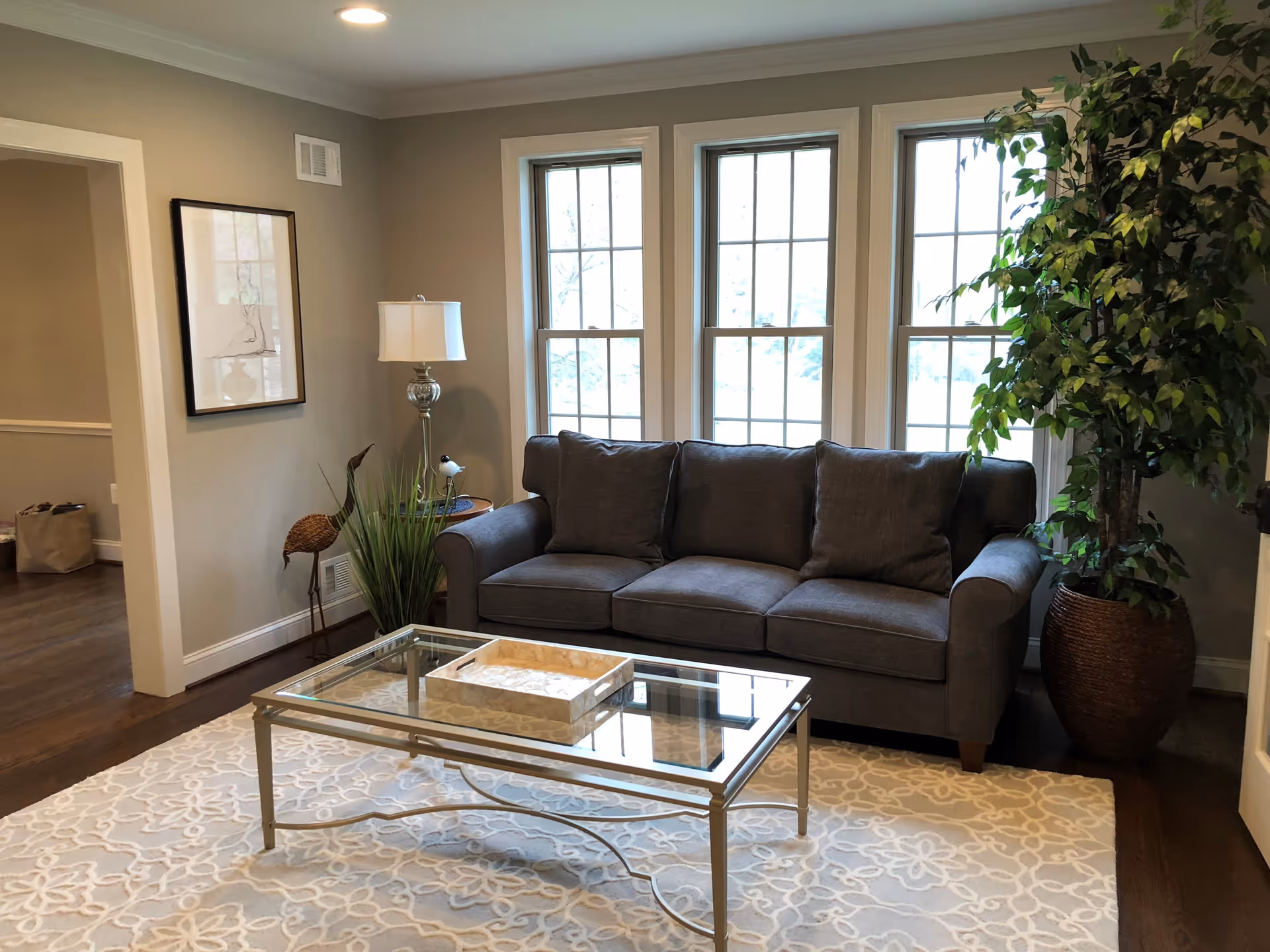 A cozy living room with a gray three-seater sofa placed in front of three large windows. A glass coffee table with a decorative tray sits on a patterned beige and white rug. To the left of the sofa is a floor lamp, a framed artwork on the wall, and a decorative bird sculpture with some green plants. On the right side, there is a large potted plant. The room has wooden flooring and beige walls.