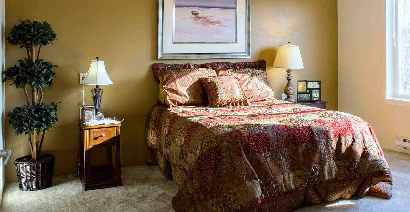 A cozy bedroom with a bed covered in a patterned quilt and matching pillows. There are two bedside tables with lamps on each side of the bed, a framed picture hanging above the bed, a potted plant in the corner, and a window letting in natural light.