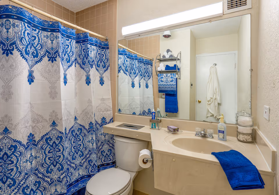 A bathroom with a beige countertop and sink, a toilet, and a shower with a blue and white patterned curtain. Above the sink is a large mirror with a light fixture. On the countertop are a blue towel, a soap dispenser, a glass container with cotton balls, and a small box. A towel rack on the wall holds rolled white towels and blue towels. A white bathrobe hangs on a hook on the door.