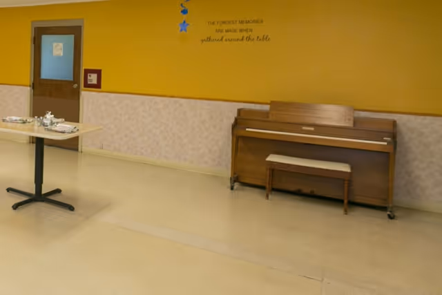 Interior room with a yellow and beige wall, a wooden upright piano with a matching bench, a table set with cups and plates, and a door with a sign on it. The wall has a quote that reads 'THE FONDEST MEMORIES ARE MADE WHEN gathered around the table'.