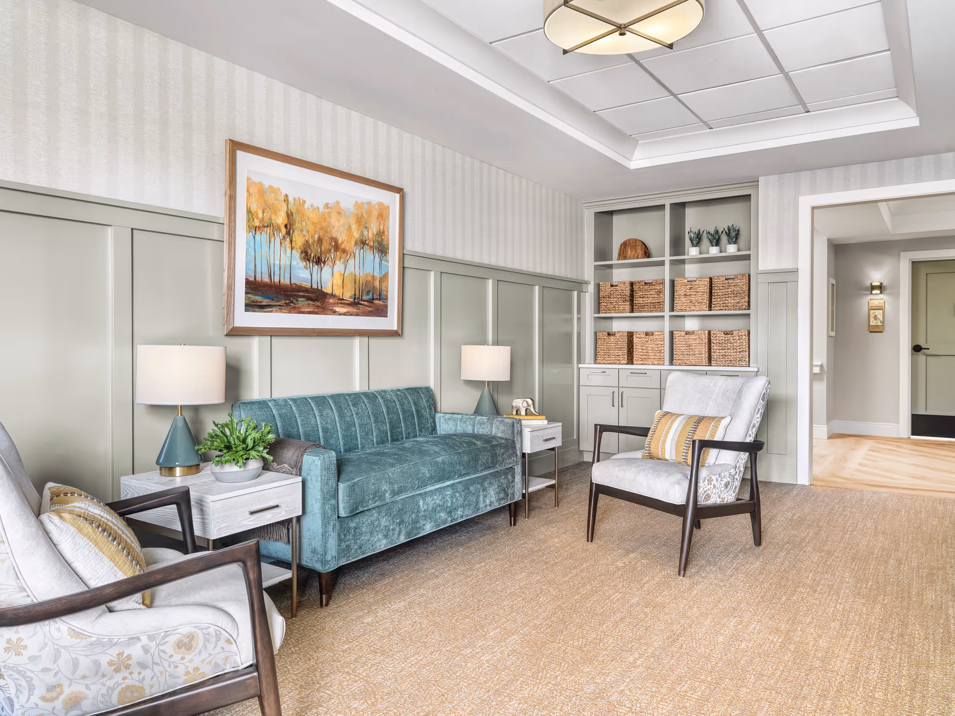 A cozy living room with a teal velvet sofa, two armchairs with patterned cushions, two side tables with lamps, a framed painting of autumn trees on the wall, built-in shelves with wicker baskets and small potted plants, and a beige carpeted floor.