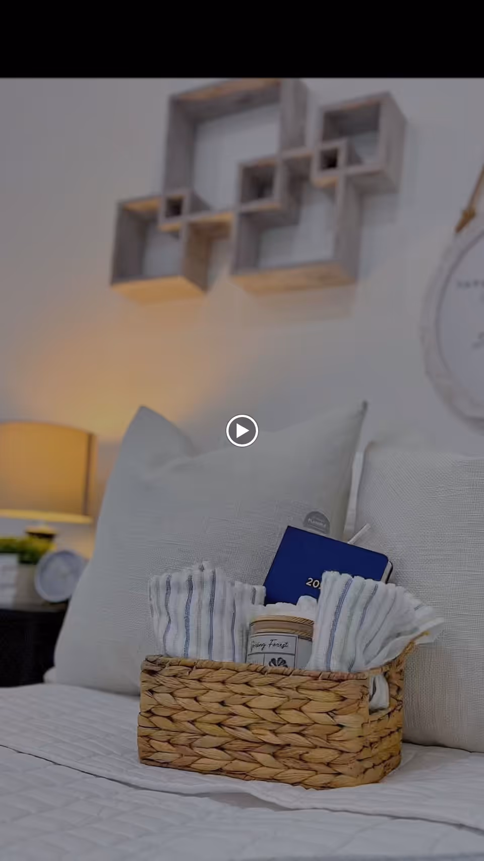 A cozy bedroom scene with a woven basket placed on a white quilted bedspread. The basket contains folded striped cloths, a small jar candle, and a blue 2023 planner. Behind the basket are two large white pillows. A bedside table with a lamp and a clock is visible in the background, along with a decorative wall shelf and a round wall hanging.