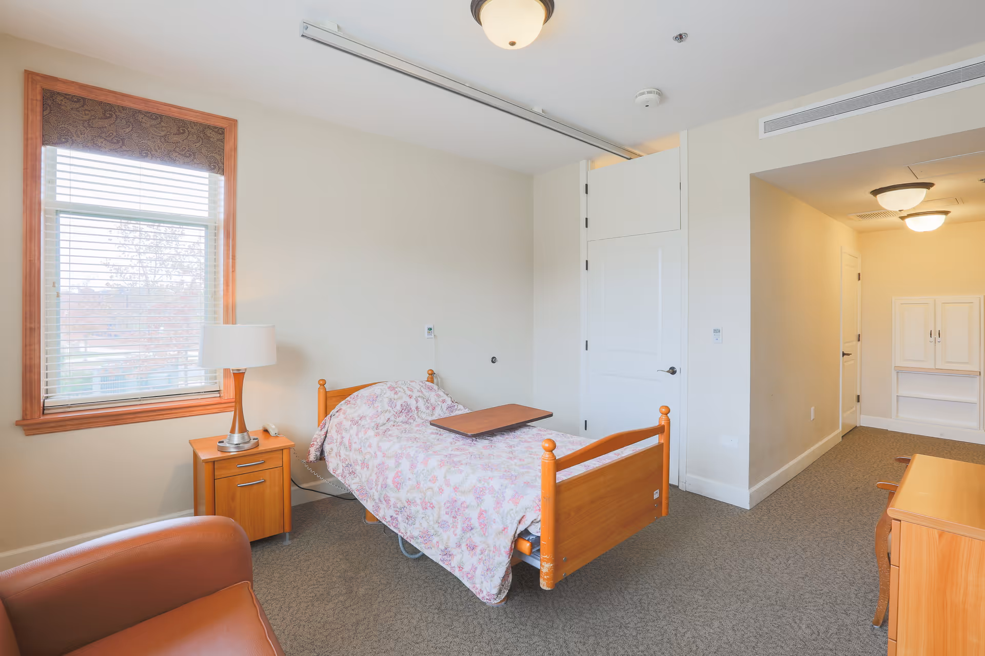 A nursing home room with a single wooden bed covered with a floral blanket and a wooden overbed table. Next to the bed is a wooden nightstand with a lamp. There is a large window with blinds and a patterned valance. A brown leather chair is partially visible in the foreground. The room has beige walls, carpeted floor, and a white door leading to a hallway with ceiling lights and a built-in cabinet.
