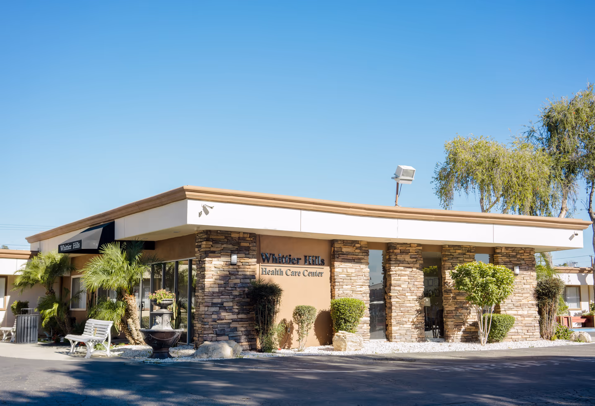 Exterior view of Whittier Hills Health Care Center building with stone pillars, windows, and landscaping including palm trees and bushes under a clear blue sky.