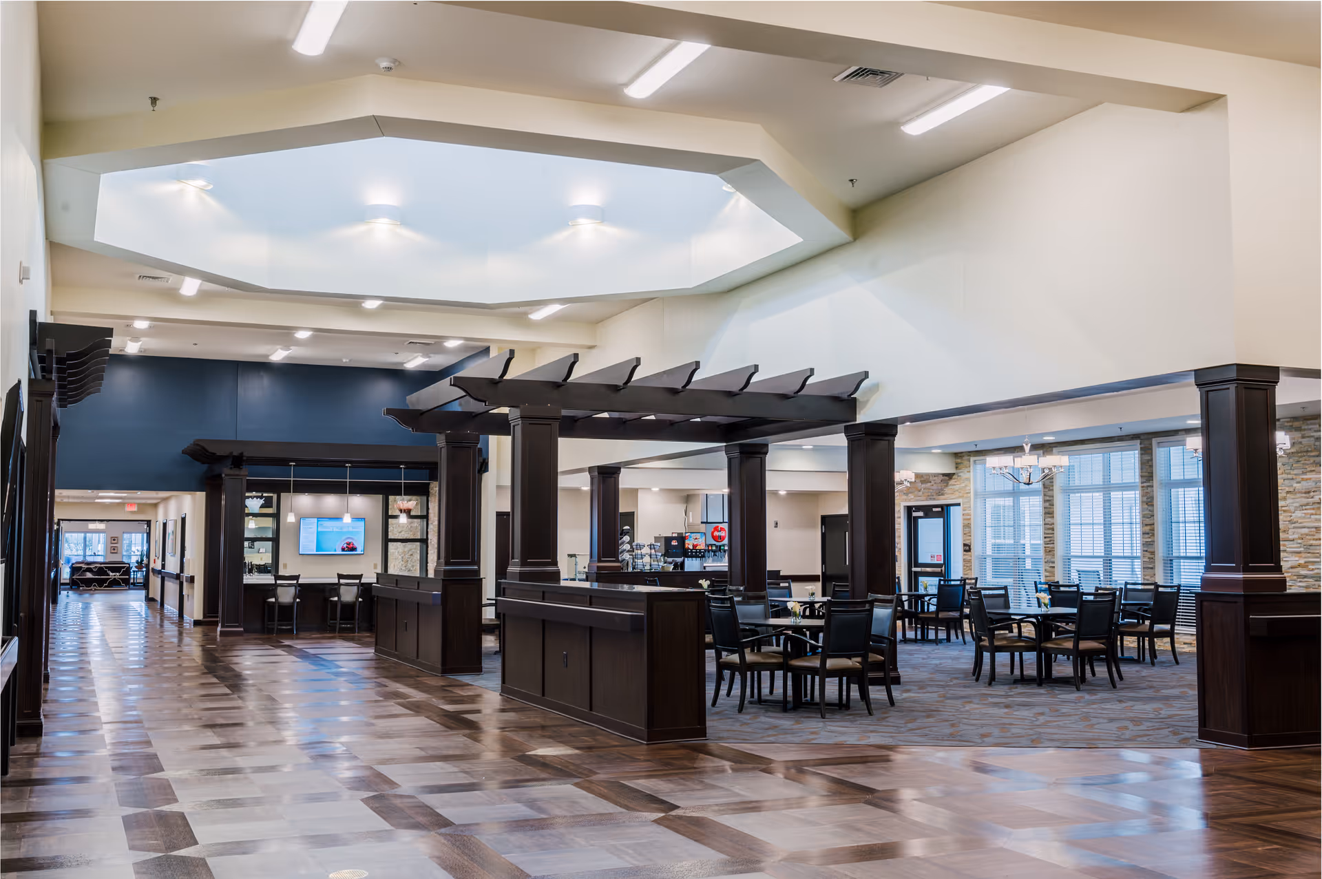 Spacious interior of a senior living facility featuring a large open area with dark wooden columns and beams, multiple tables and chairs arranged for dining or socializing, a high ceiling with a skylight, and a seating area with a TV in the background.
