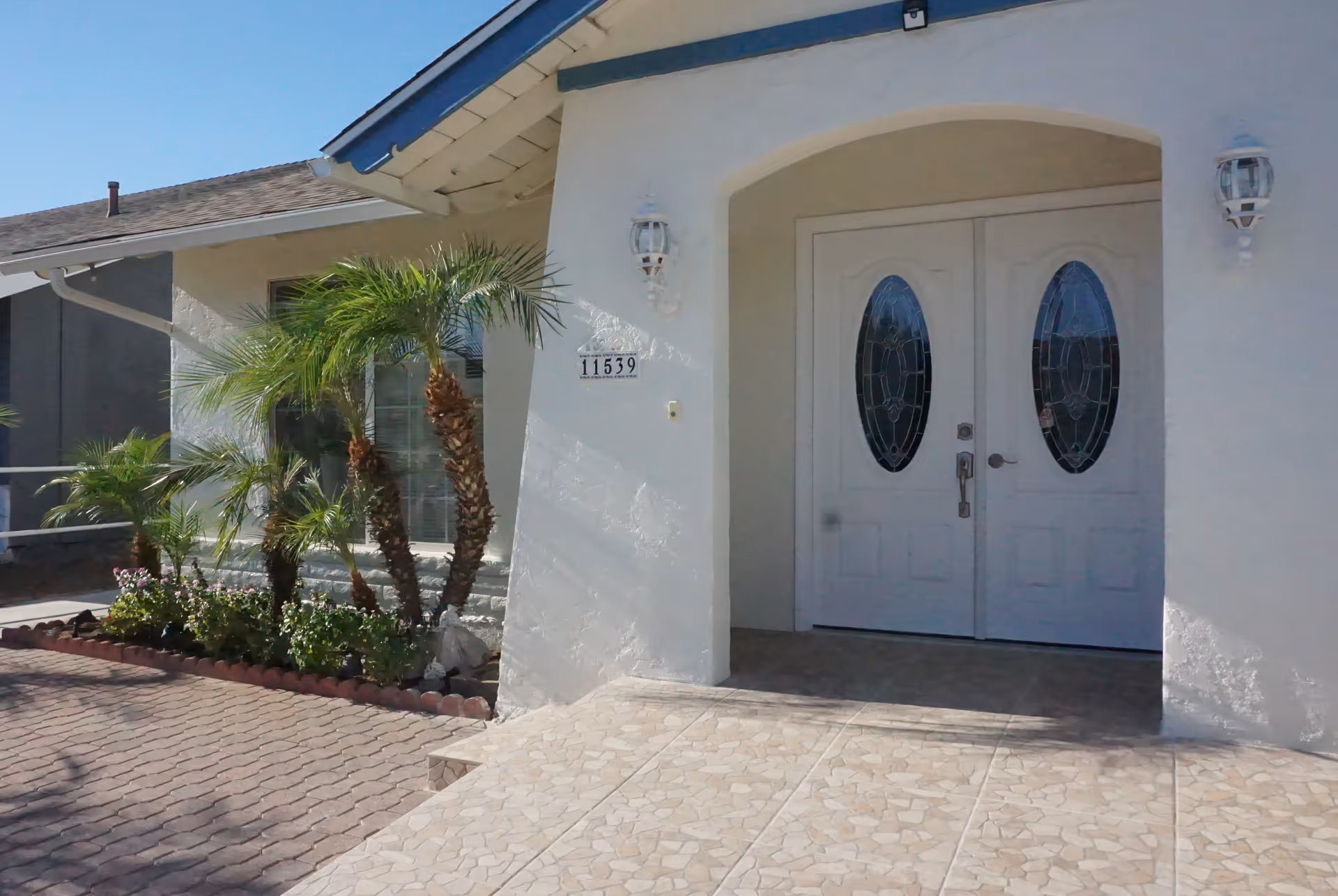 Front entrance of a white building with double doors featuring oval glass panels, a tiled porch, and small palm trees and plants along the side.