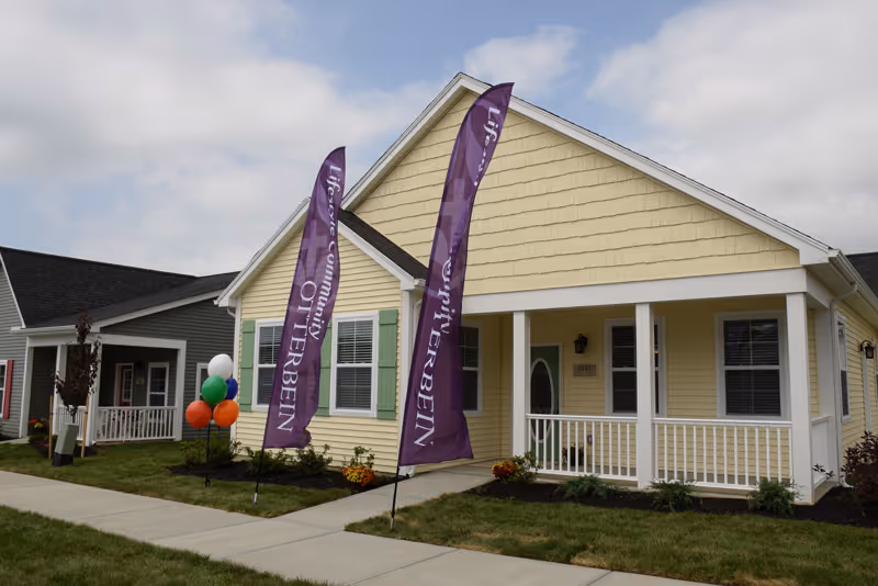 Exterior view of a single-story yellow house with a front porch and white railing. Two tall purple flags with the text 'Otterbein' are placed in the front yard, along with a cluster of colorful balloons. Another similar house painted gray is visible next to it under a partly cloudy sky.
