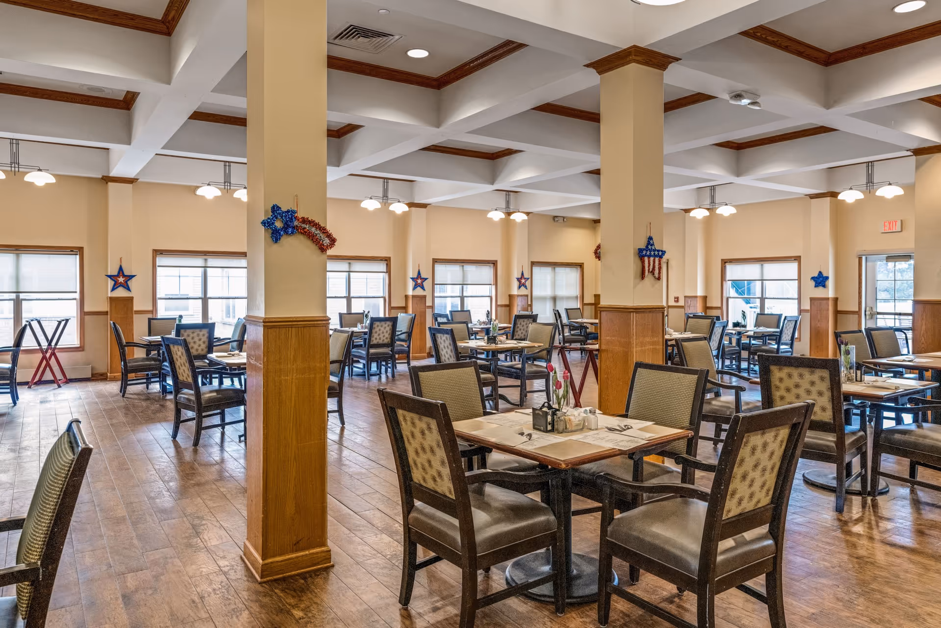 A spacious dining room with multiple tables and chairs arranged neatly. The room features wooden flooring, beige walls with wooden paneling, and large windows allowing natural light to enter. The ceiling has a grid pattern with recessed lighting and hanging lamps. Patriotic star decorations are hung on the columns and walls.