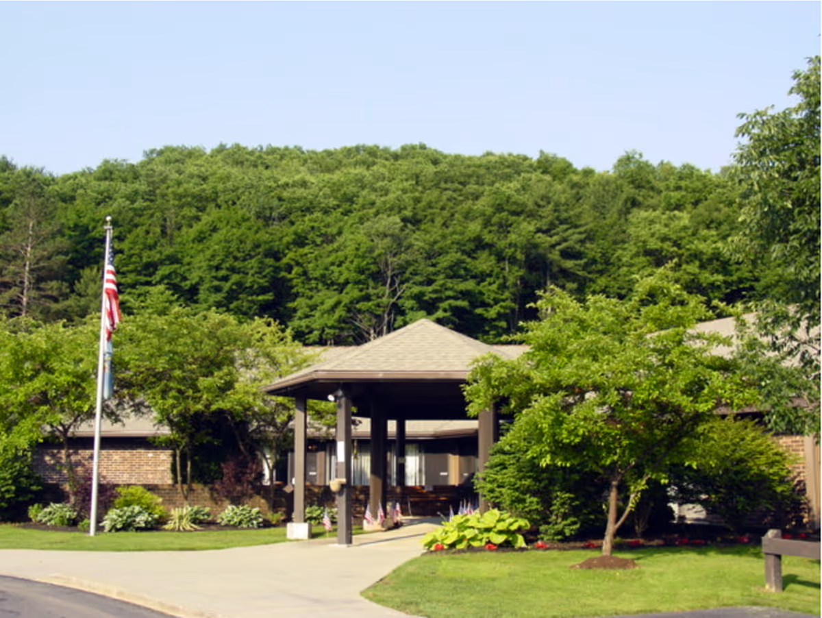 Front entrance of a single-story brick building with a covered driveway, an American flag, and landscaped trees and lawn in front of a wooded hillside.