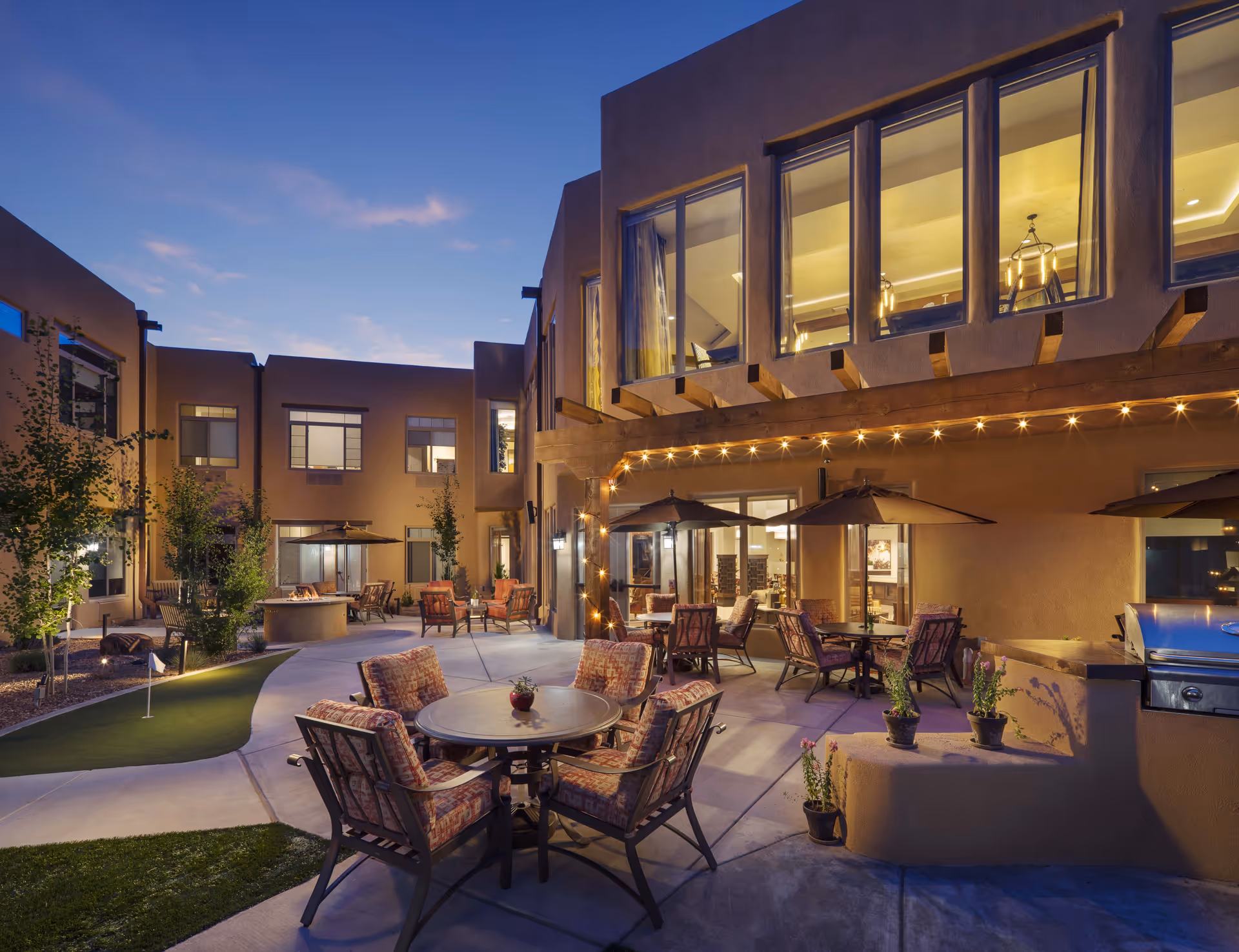 Outdoor courtyard area of MorningStar Assisted Living & Memory Care of Santa Fe at dusk, featuring patio tables with cushioned chairs and umbrellas, string lights hanging from the building, potted plants, a putting green, and a built-in grill. The building has large windows and a southwestern architectural style.