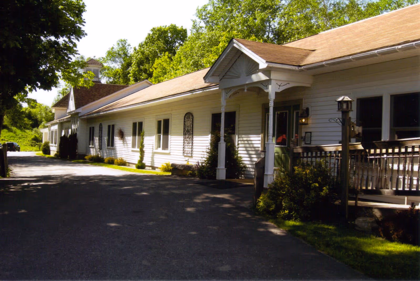 Exterior view of a single-story white building with a brown roof, surrounded by greenery and trees. The building has multiple windows, a covered entrance with decorative columns, and a paved driveway leading up to it.