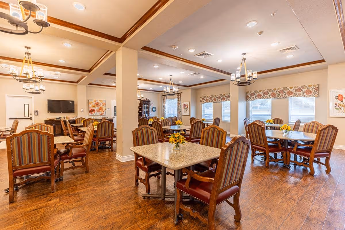 Bright dining room with multiple tables and upholstered chairs on wood floors under chandeliers.