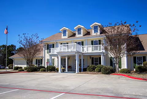 Front exterior view of a two-story senior living facility building with a beige facade, white trim, and a covered entrance supported by white columns. There are three dormer windows on the roof, small trees and bushes around the building, an American flag on a flagpole to the left, and a clear blue sky above.