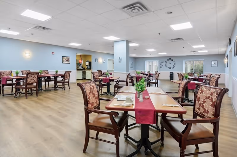 A spacious dining room in a senior living facility with multiple wooden tables and cushioned chairs arranged neatly. Each table has a red table runner, a small green plant centerpiece, and place settings with napkins and cups. The room has light blue walls, large windows with vertical blinds, and a clock on the far wall.
