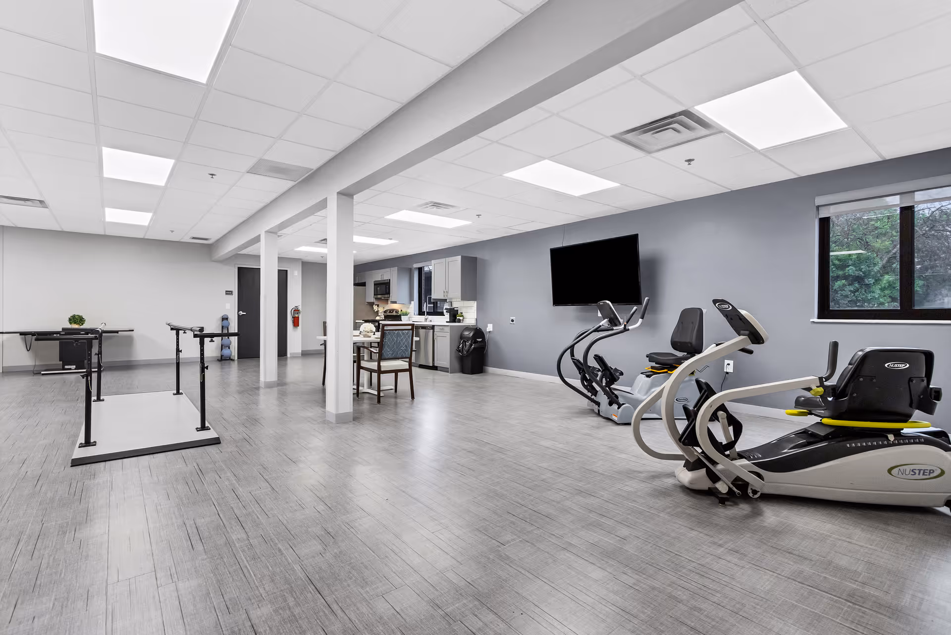 A spacious rehabilitation or exercise room with parallel bars for walking practice, two recumbent exercise bikes, a wall-mounted TV, a small table with chairs, and a kitchenette area in the background. The room has large windows, gray walls, and a light gray wood-patterned floor.