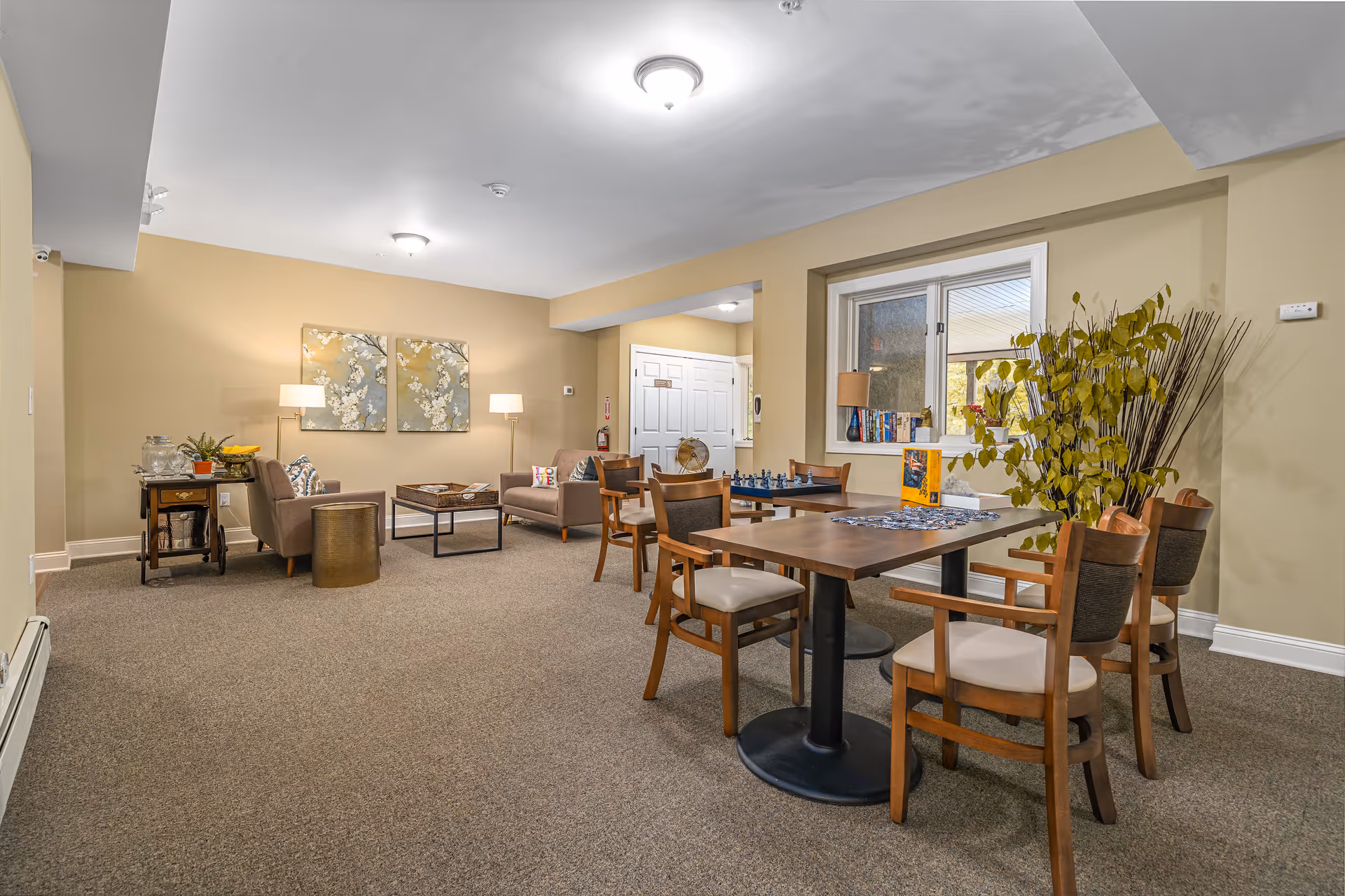 A cozy common area in The Lambertville Estate featuring two wooden tables with chairs, a chess set, and a puzzle on the tables. There are two armchairs with a small coffee table between them, two floor lamps, and decorative wall art. A window with a small lamp and books on the sill is visible, along with a potted plant and a small side table with a water dispenser and fruit bowl.