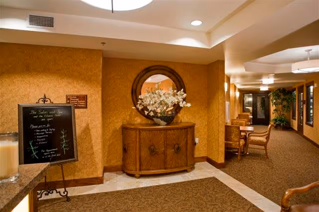 Interior hallway of a retirement community with warm yellow walls, a wooden cabinet with a round mirror above it, a vase with white flowers on the cabinet, a decorative sign on a stand, and seating area with chairs and plants further down the hallway.