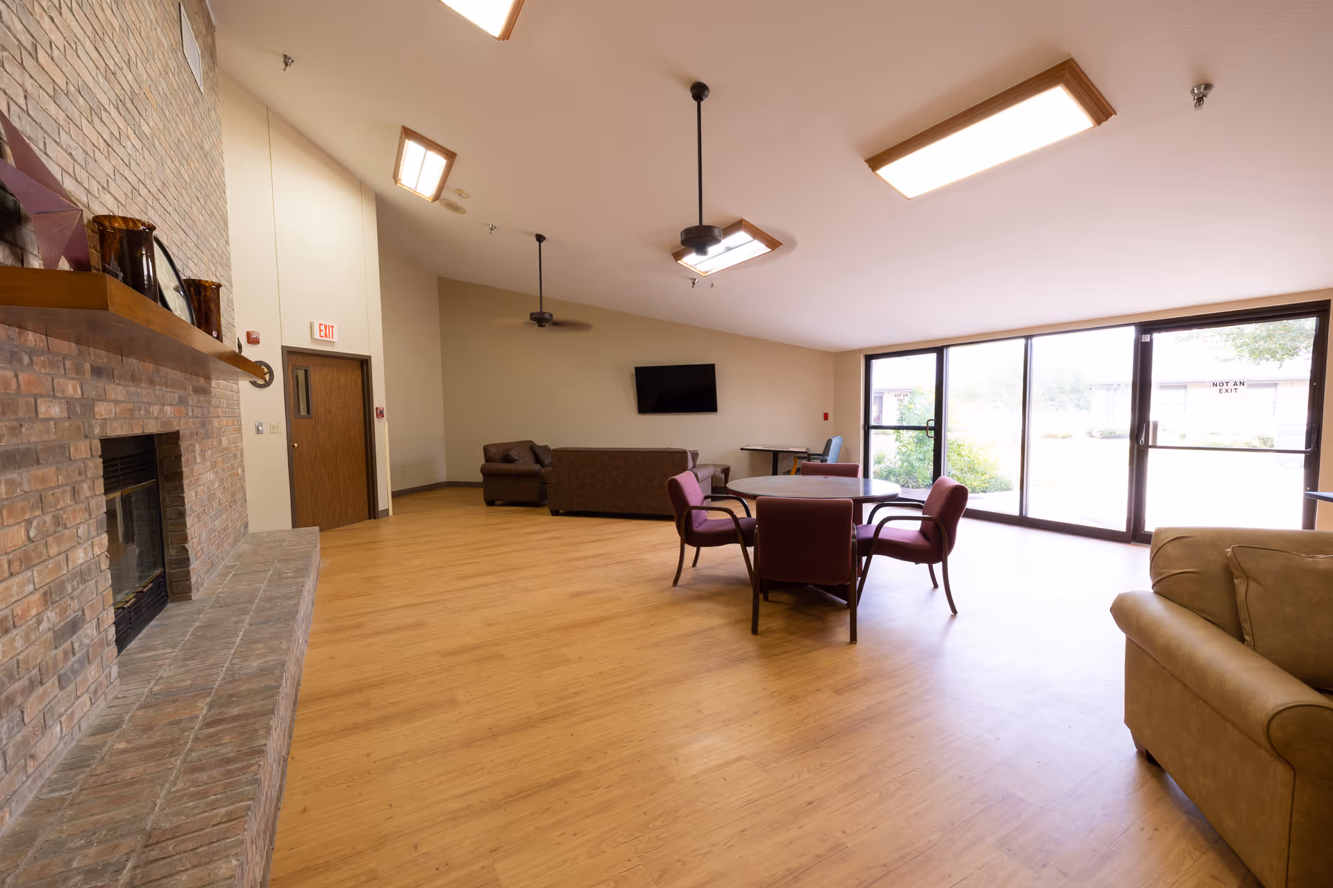 Spacious common area with wooden flooring, a brick fireplace on the left, several chairs and tables arranged in the center, a wall-mounted TV, large glass sliding doors letting in natural light, and ceiling fans.