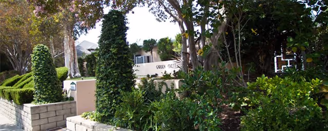 Outdoor garden area with lush green plants, trees, and trimmed bushes surrounding a sign that reads 'Garden Crest'.