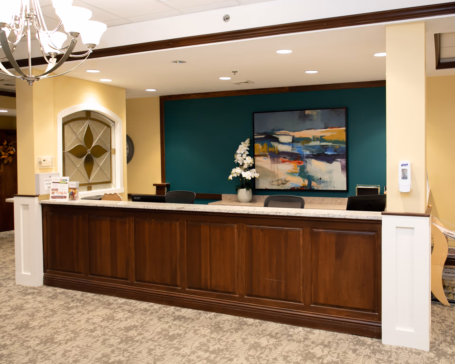 Reception desk area with a wooden front counter, two office chairs behind the desk, a vase with white flowers, and an abstract painting on a teal accent wall. The surrounding walls are painted yellow, and there is a chandelier hanging from the ceiling.
