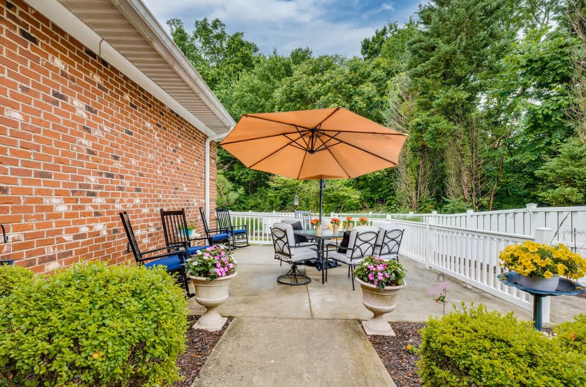 Outdoor patio area at Kerner Ridge Assisted Living featuring a large orange umbrella over a round table with four cushioned chairs. The patio is surrounded by a white fence, with potted flowers and green bushes along the concrete walkway. A brick building wall with several black rocking chairs is visible on the left side, and trees and greenery are in the background under a partly cloudy sky.