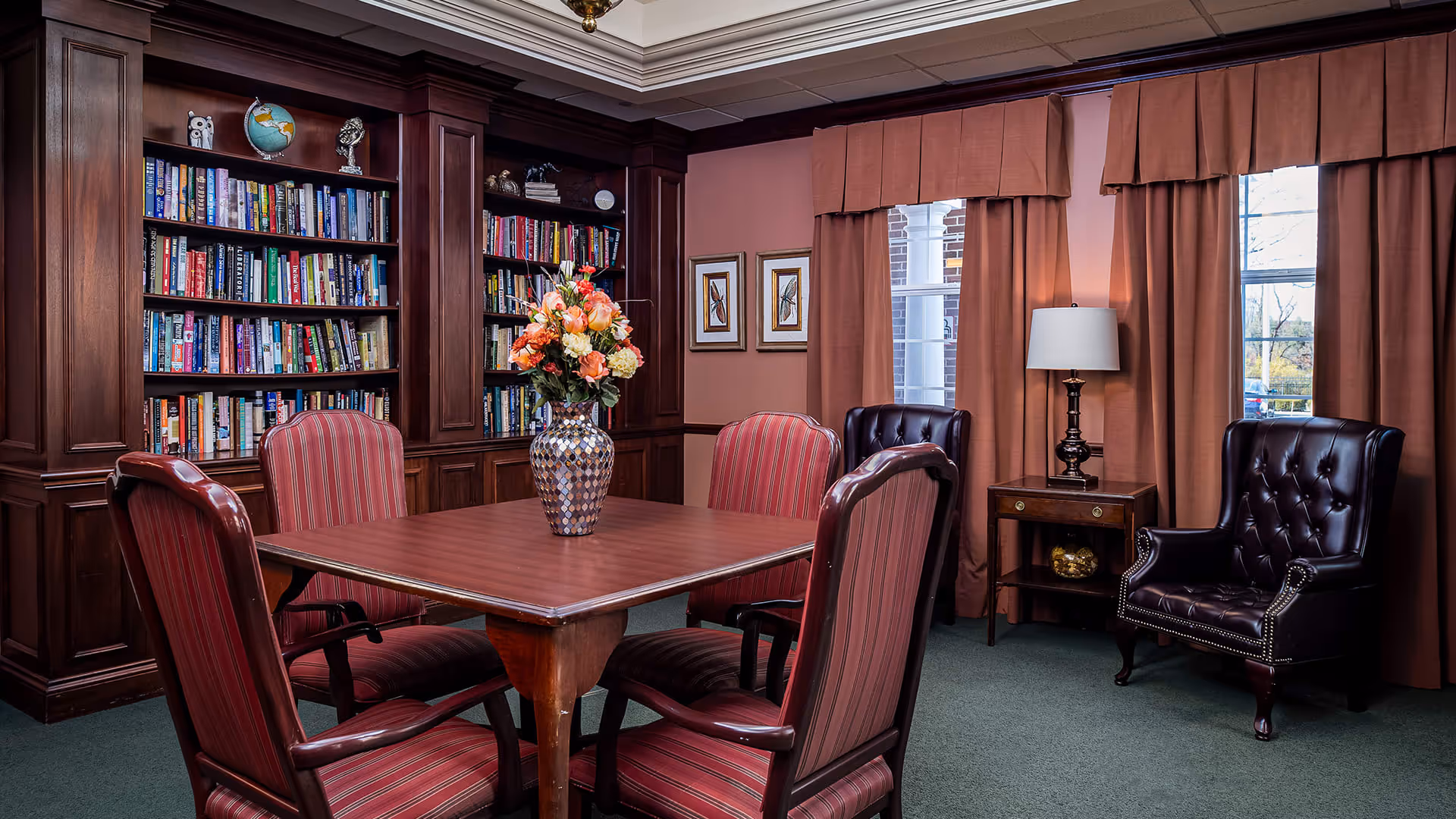 A cozy interior room with a square wooden table surrounded by four red-striped upholstered chairs. Behind the table is a built-in wooden bookshelf filled with books and decorative items. To the right, there are two dark leather armchairs, a wooden side table with a lamp, and two windows with brown curtains. A vase with a bouquet of flowers is placed on the table.