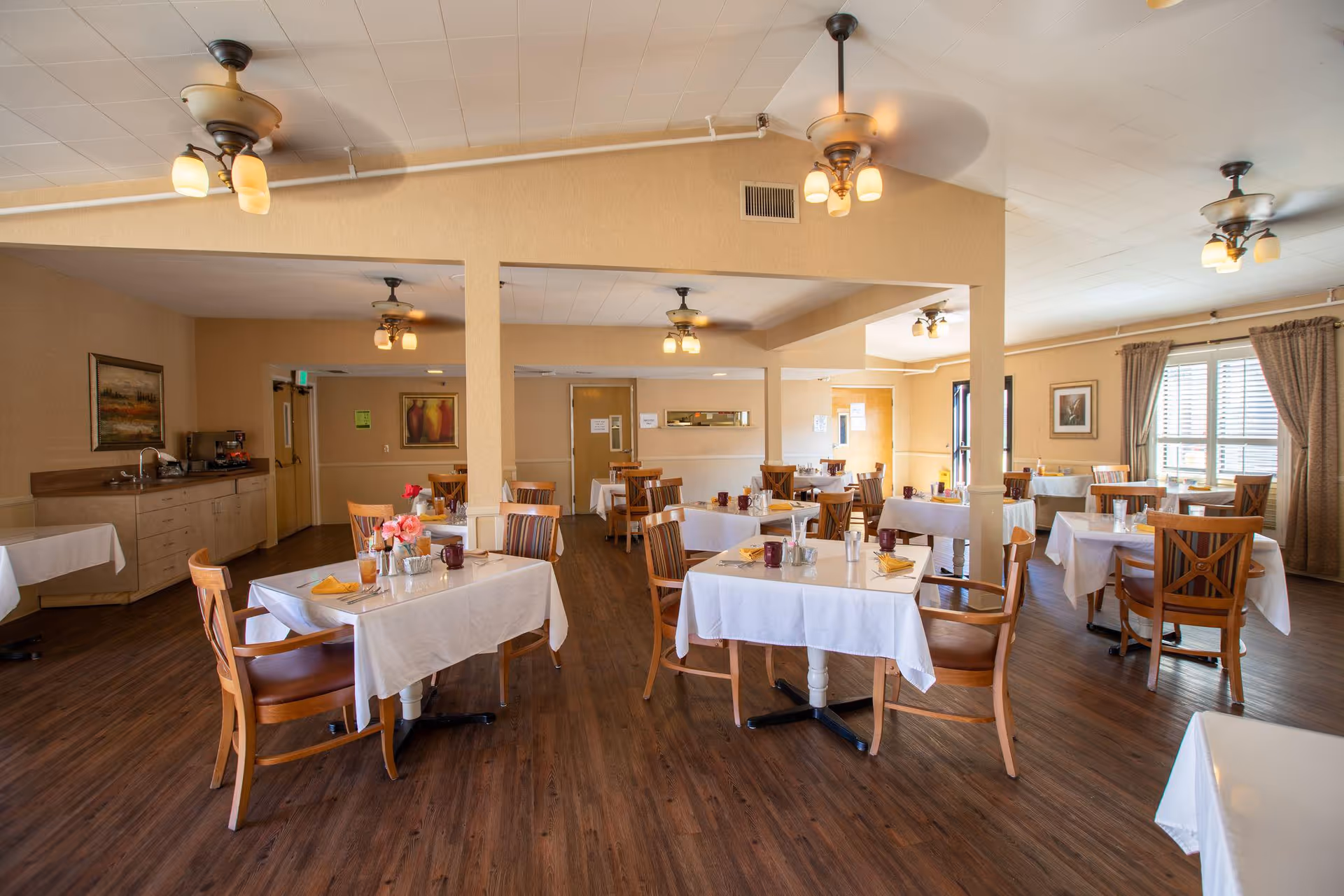 A bright and spacious dining room with multiple tables covered in white tablecloths, each set with napkins, cups, and small flower arrangements. Wooden chairs surround the tables, and the room features wood flooring, beige walls, ceiling fans with lights, and large windows with curtains allowing natural light to enter.