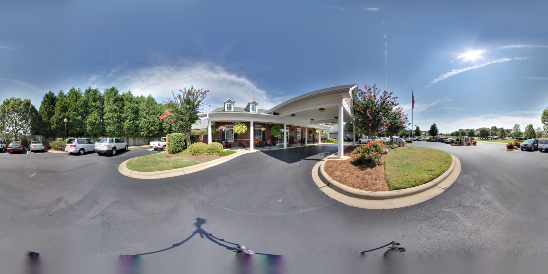 Wide panoramic view of the front exterior of Summit Place of Mooresville, showing a covered entrance with white columns, a brick building, landscaped bushes and trees, a flagpole with an American flag, and a parking lot with several cars under a clear blue sky.