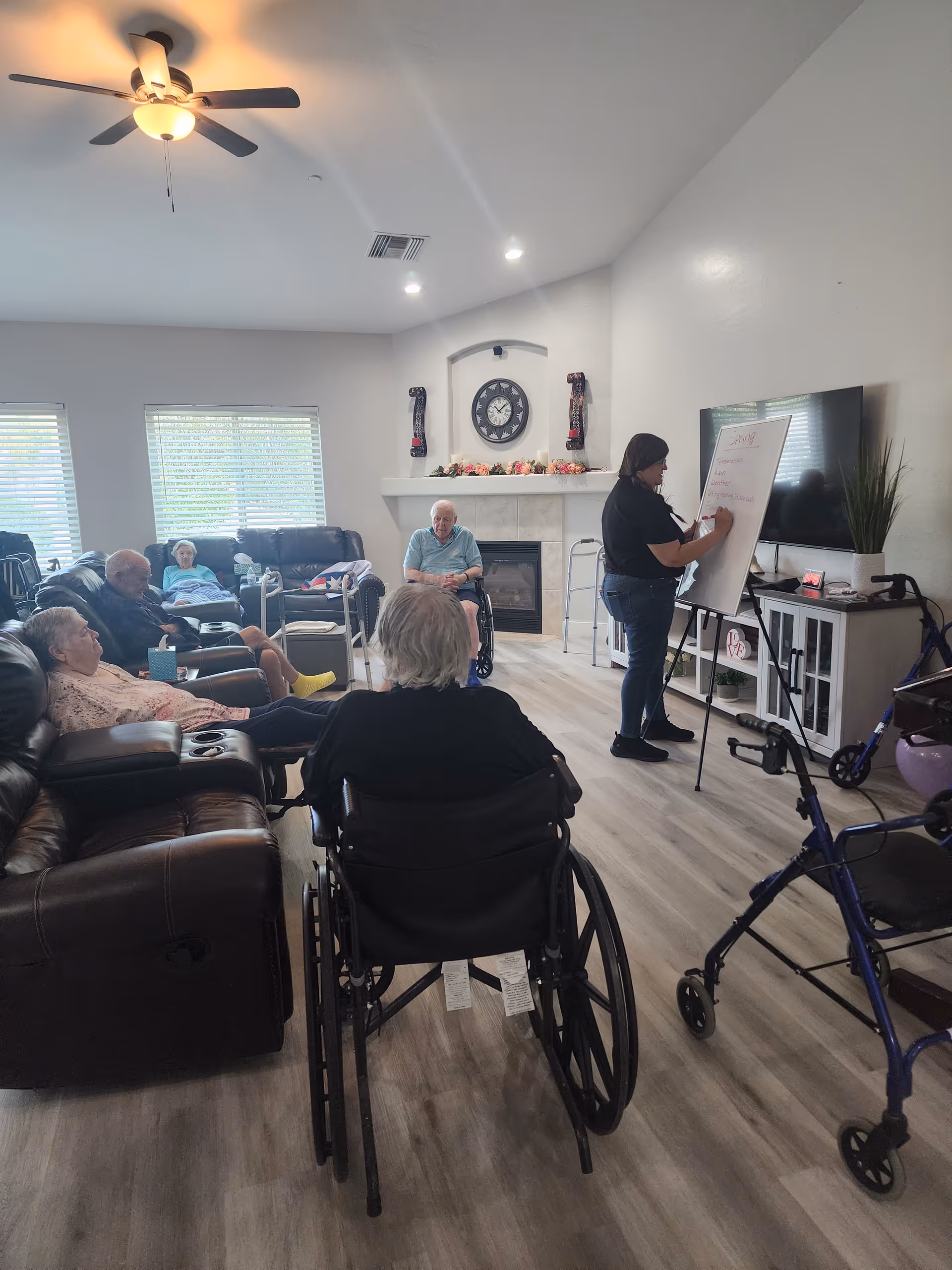 A group of elderly people seated in a living room area, some in wheelchairs and others in recliners, attentively watching a woman who is writing on a whiteboard. The room has a ceiling fan, a fireplace with a clock above it, and a large TV on a stand with some plants and decorations.