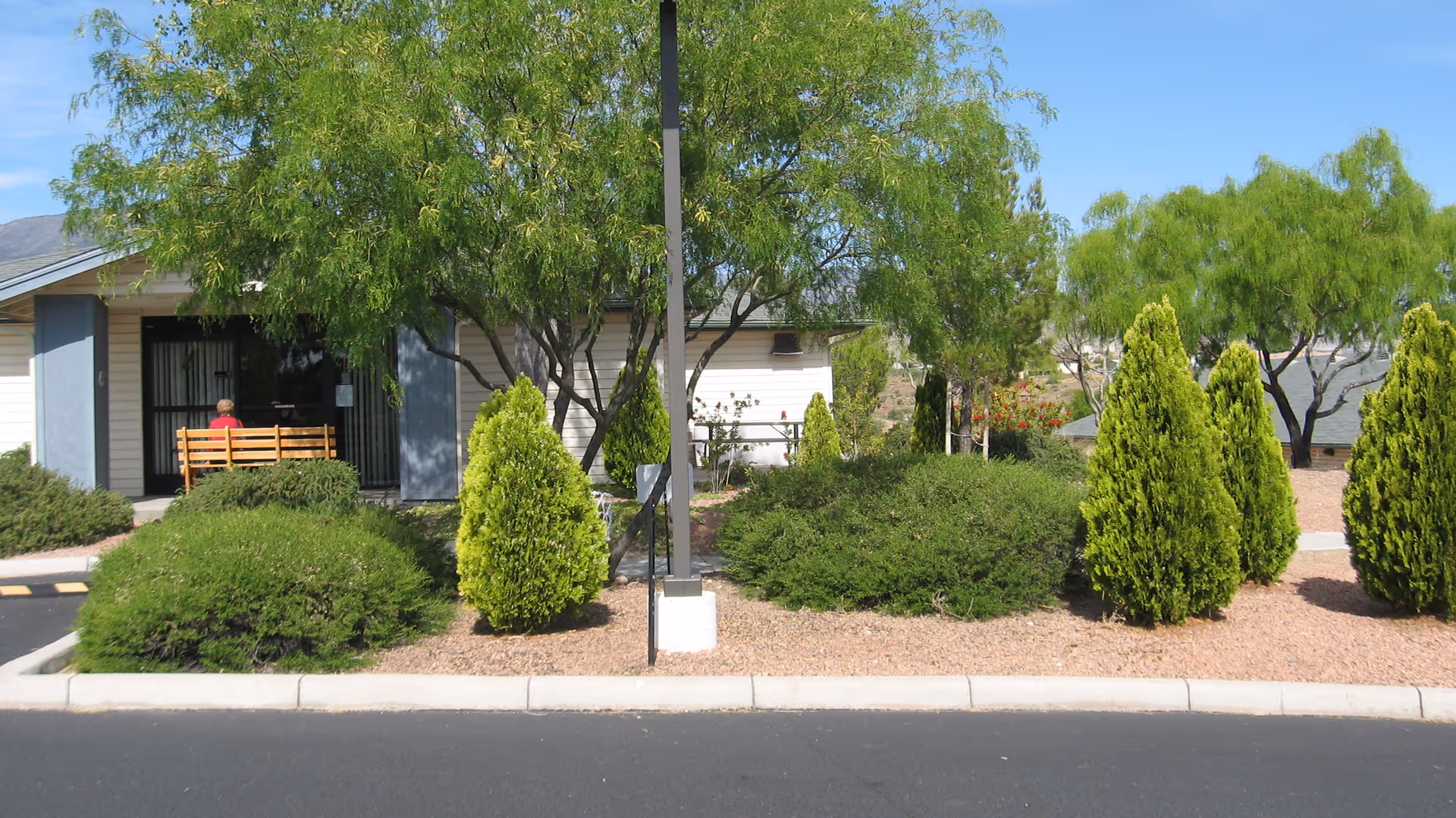 View of a single-story building with light-colored siding and a small porch area with a bench. The foreground features a landscaped area with various green bushes and trees, and a paved road in front. A person wearing a red shirt is sitting on the bench near the building entrance.