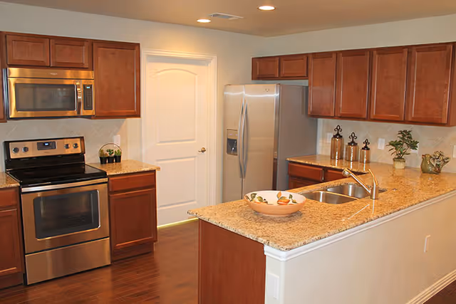 Modern kitchen with wooden cabinets, stainless steel appliances including a microwave, stove, and refrigerator, granite countertops, a double sink, and decorative items on the counter.