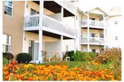 Exterior view of a senior housing apartment building with multiple balconies and a garden filled with orange and yellow flowers in the foreground.