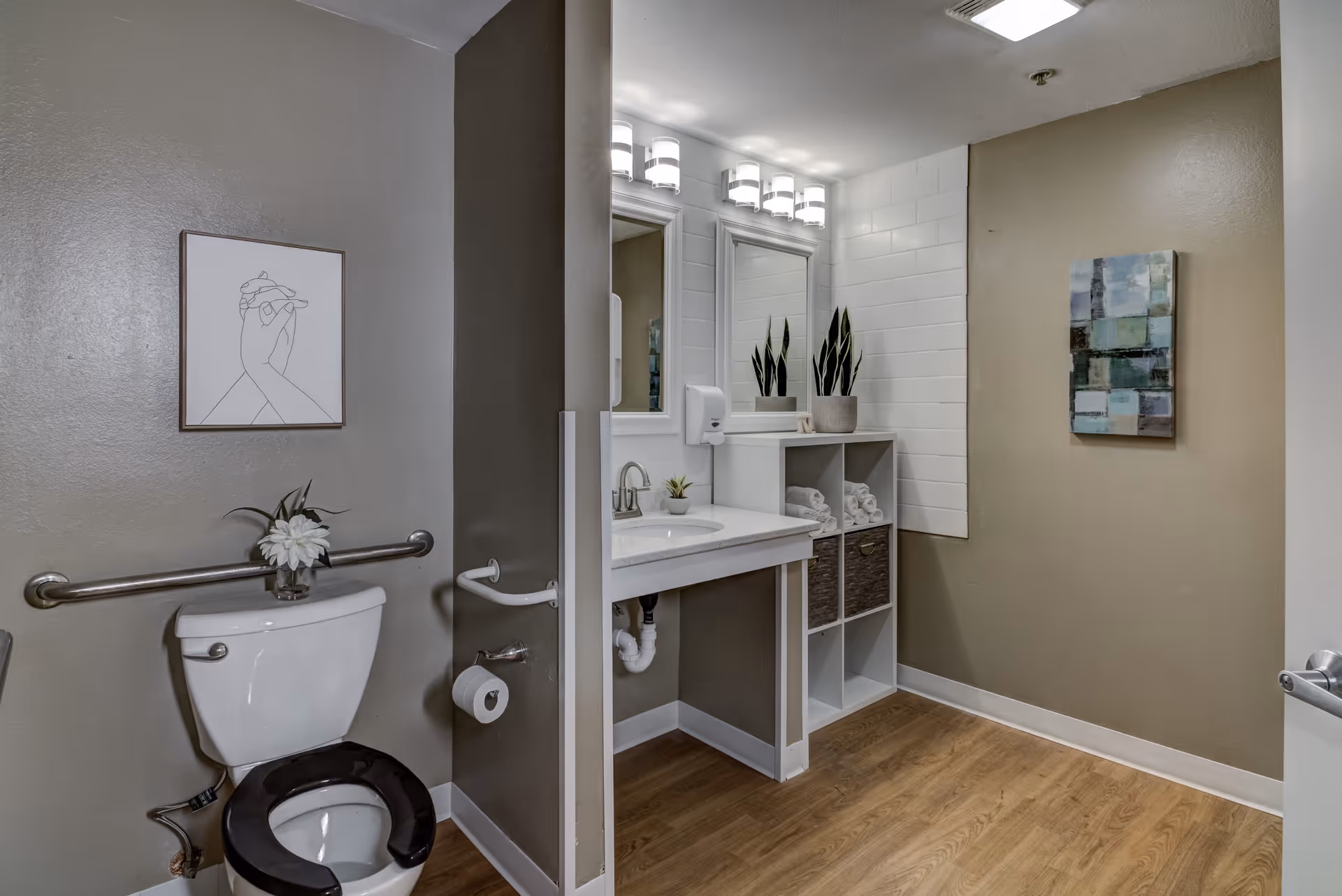 A clean and modern bathroom with a white toilet featuring a black seat, a grab bar on the wall, and a small vase with a white flower on the toilet tank. The bathroom has a white sink with a countertop, two mirrors above the sink, and a white shelving unit holding rolled towels and baskets. The walls are painted beige, and there is a piece of abstract artwork on the wall. The floor is wood-style laminate.