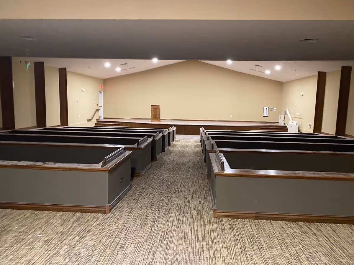 Interior view of a room with multiple rows of wooden pews facing a small stage with a wooden podium. The walls are beige, and the carpet is patterned in neutral tones. The room is well-lit with ceiling lights.