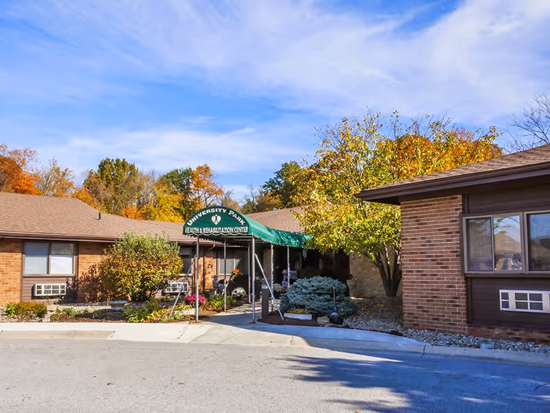 Exterior view of University Park Rehabilitation and Healthcare Center showing a single-story brick building with a green awning over the entrance. The surrounding area has trees with autumn foliage and some landscaping with bushes and flowers.