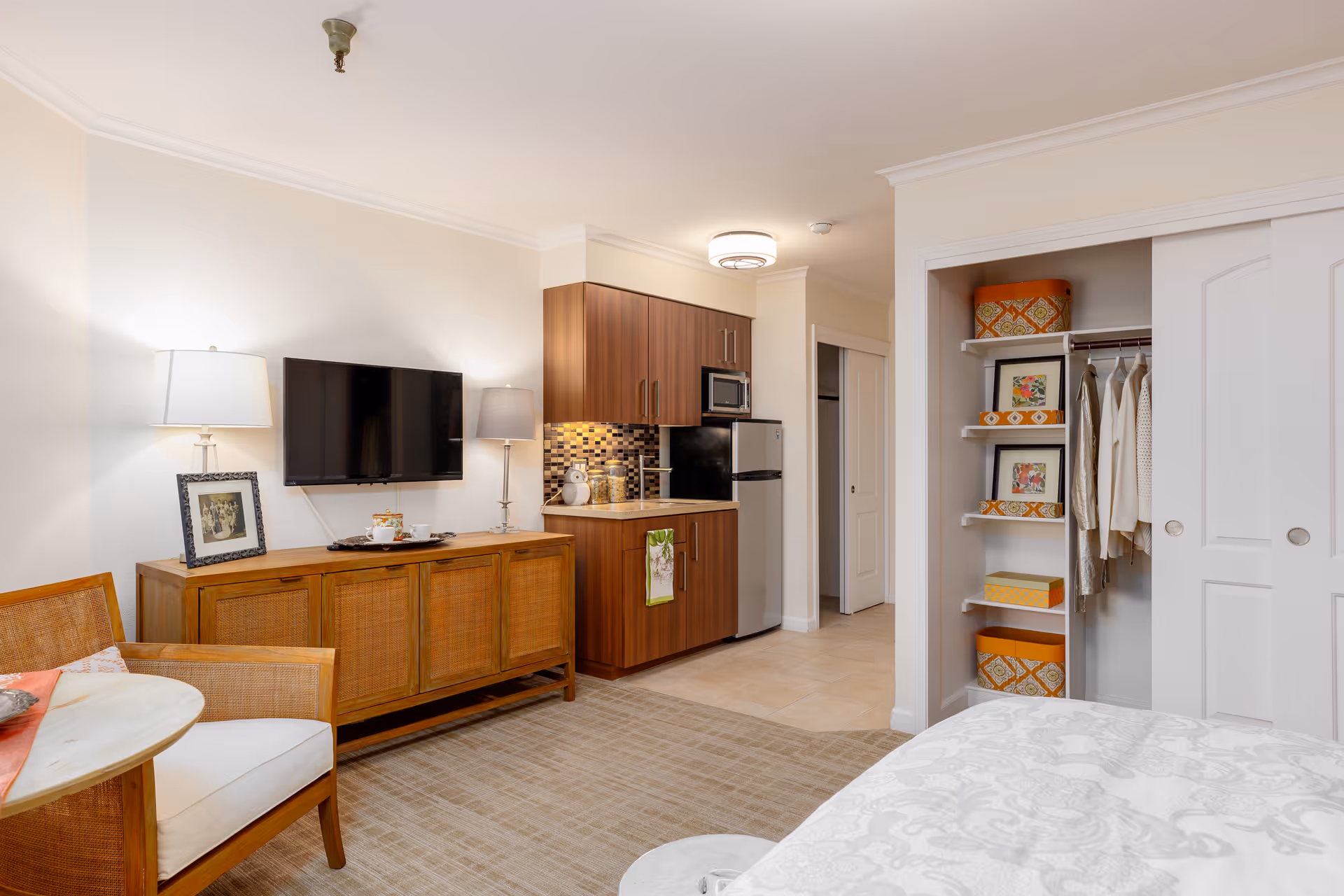 Bright studio apartment interior with a kitchenette, wall-mounted TV on a wooden console, seating area, and an open closet next to a bed.