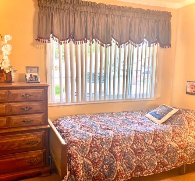 Bedroom in a senior living facility with a quilted bedspread, dresser, photos, and blinds
