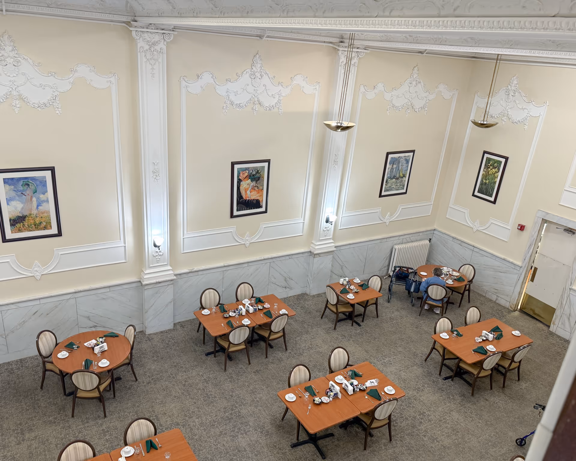 A dining room with several wooden tables and chairs arranged neatly. Each table is set with white plates, cups, silverware, and green napkins. The walls are cream-colored with ornate white molding and framed paintings. There are two pendant lights hanging from the ceiling. An elderly person is seated at one of the tables near the corner with a walker beside them.