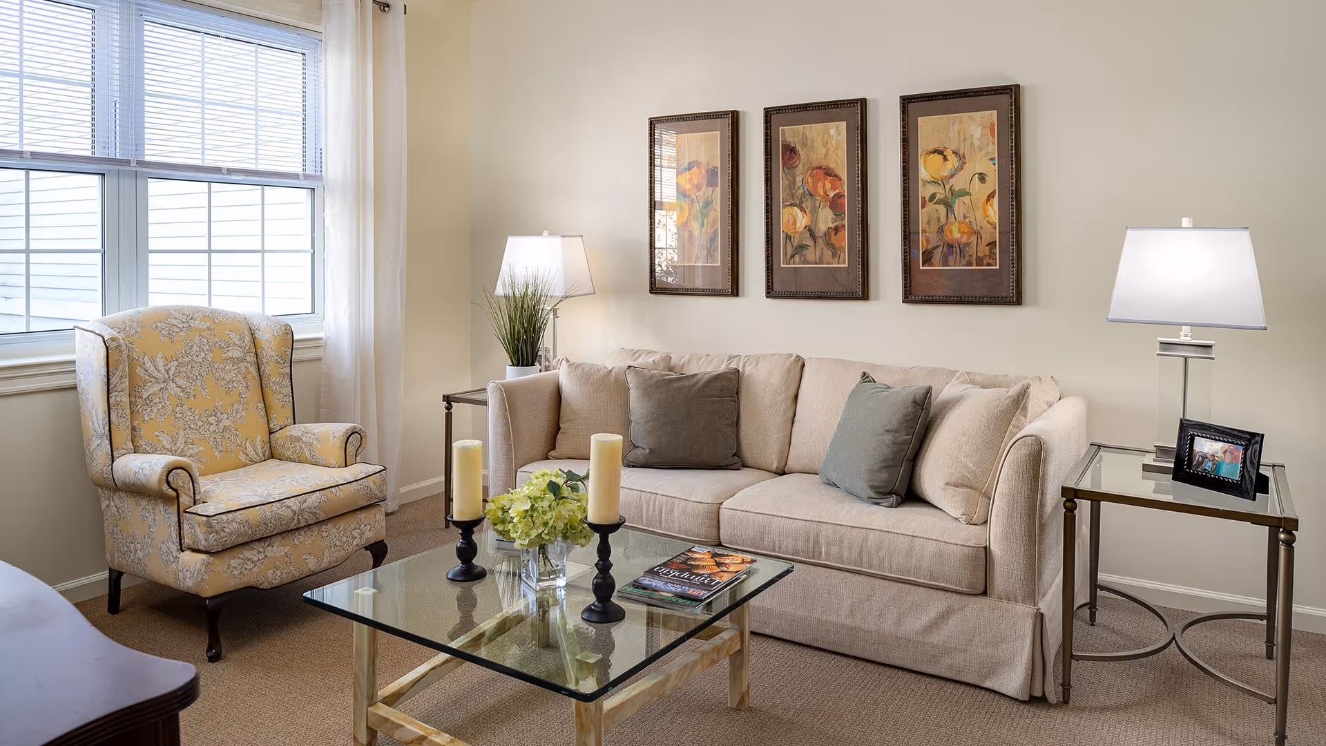 A cozy living room with a beige sofa adorned with multiple cushions, a glass coffee table with candles and a flower arrangement, a yellow patterned armchair near a window with white curtains, two side tables each with a lamp, and three framed floral paintings on the wall.