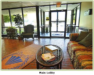 Main lobby area with a glass entrance door, a patterned rug on the brick floor, a glass coffee table with magazines, a cushioned armchair, and a sofa with striped upholstery and pillows.