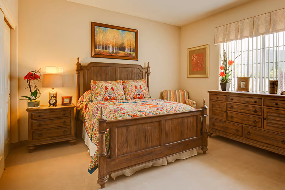 Sunlit bedroom with a wooden bed and colorful bedding, matching nightstand and dresser by a window.