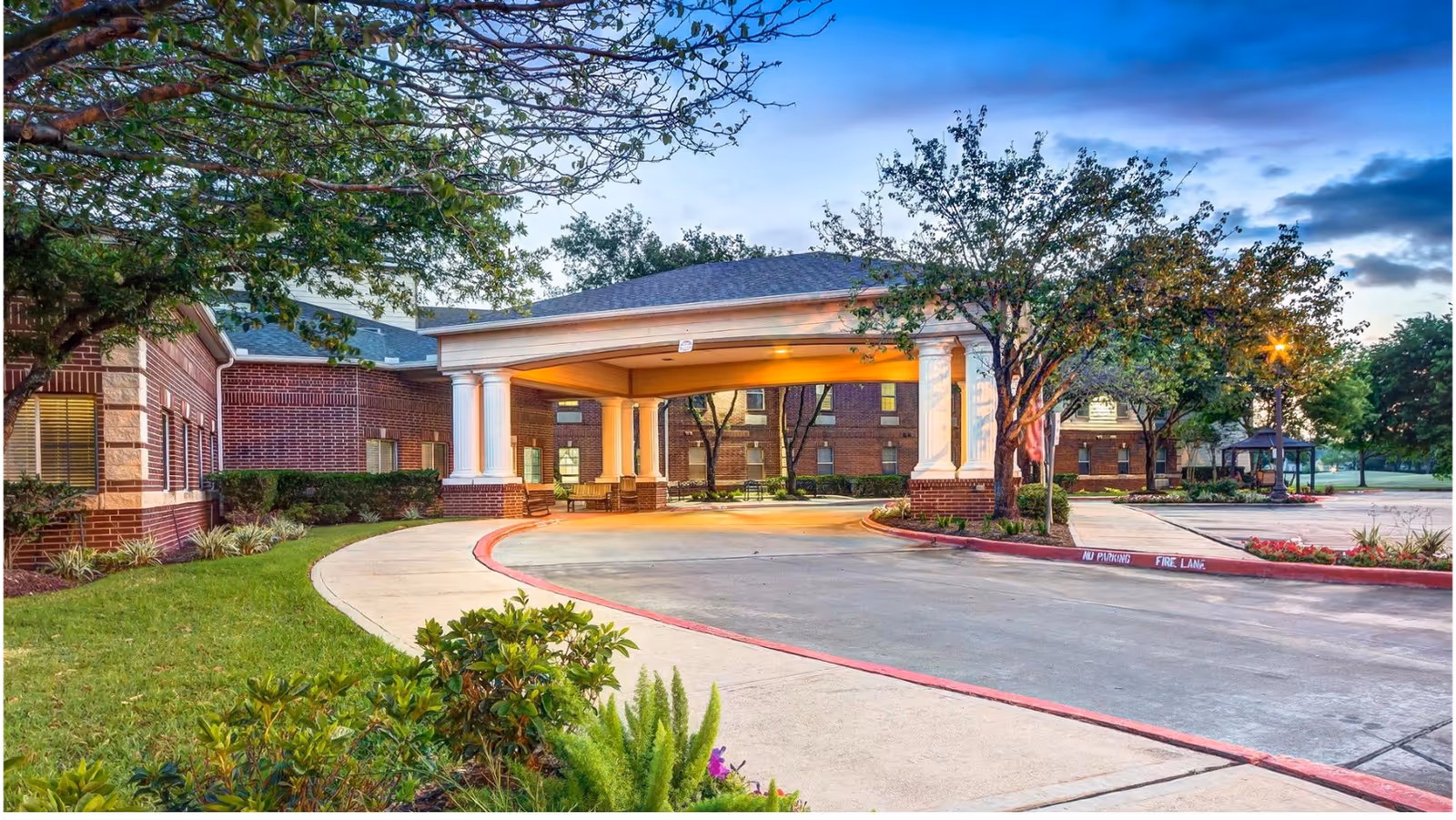 Covered driveway and entrance (porte-cochere) of a brick senior living facility with landscaped grounds and trees at dusk.